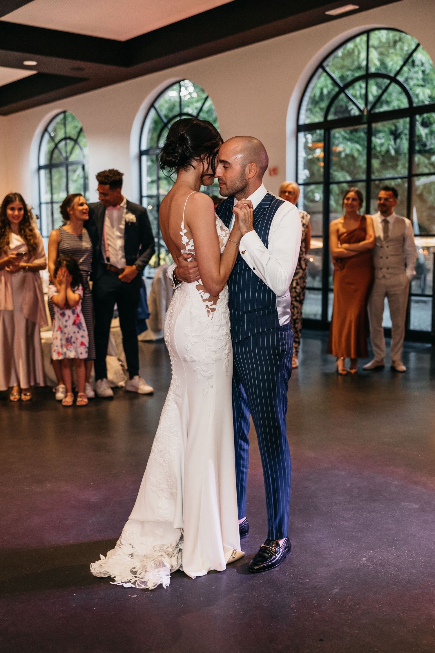 Bride and groom sharing intimate first dance in elegant venue with arched windows and wedding guests watching.