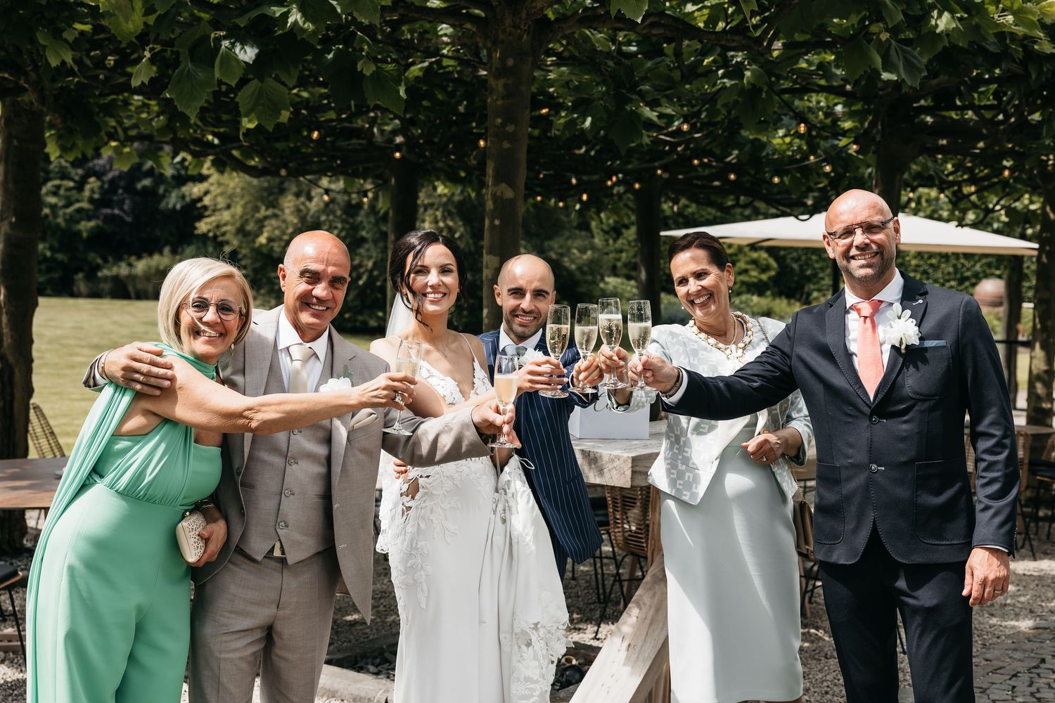 Wedding party of six toasting champagne glasses outdoors with lush green trees in background.