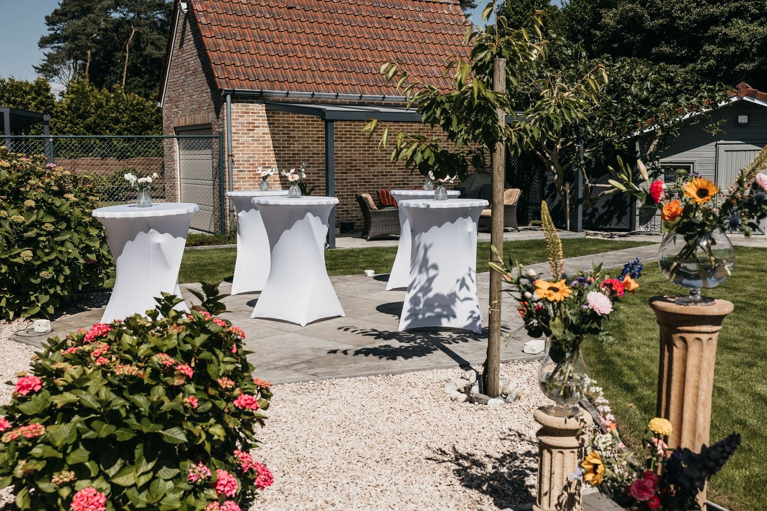 Outdoor wedding reception setup with white cocktail tables and floral decorations in a garden.