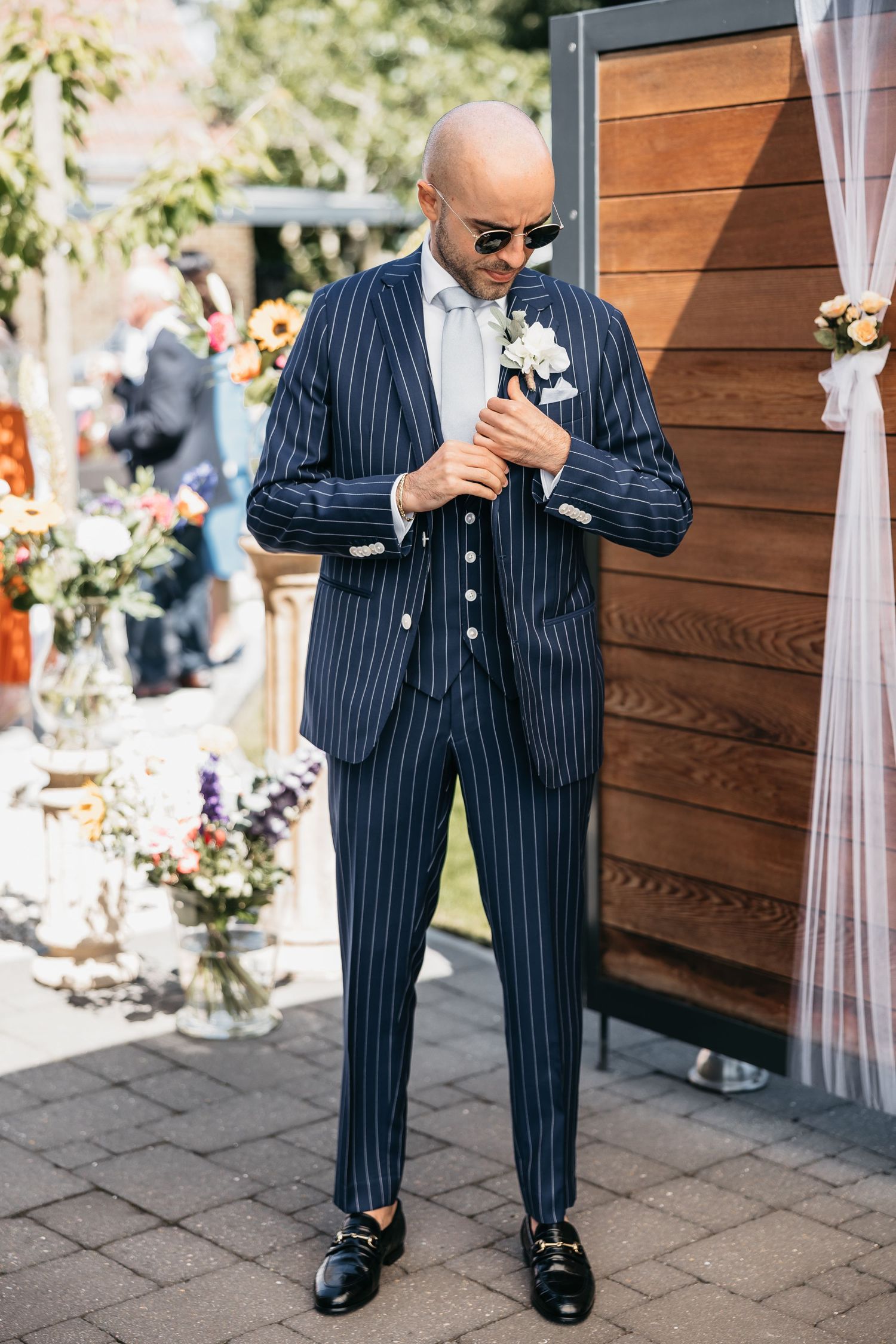 Bald groom in navy pinstripe three-piece suit with boutonniere and sunglasses at outdoor wedding.