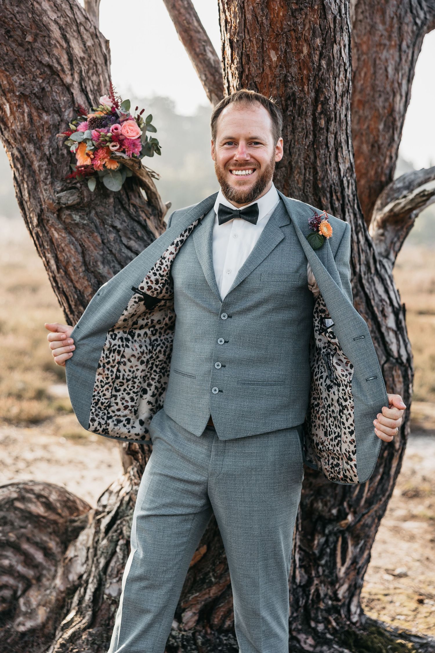 Groom in grey suit proudly displays leopard print jacket lining while standing against a pine tree outdoors.