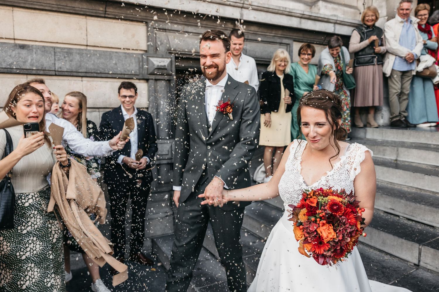 Bearded groom and smiling bride walk through confetti shower surrounded by celebrating wedding guests.
