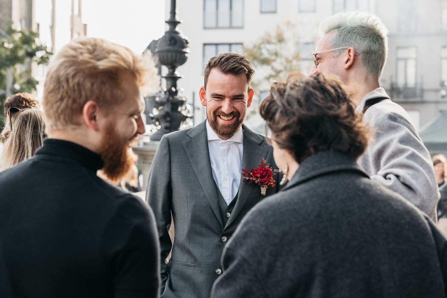 Smiling groom in grey suit with red boutonniere laughing with guests at outdoor wedding celebration.