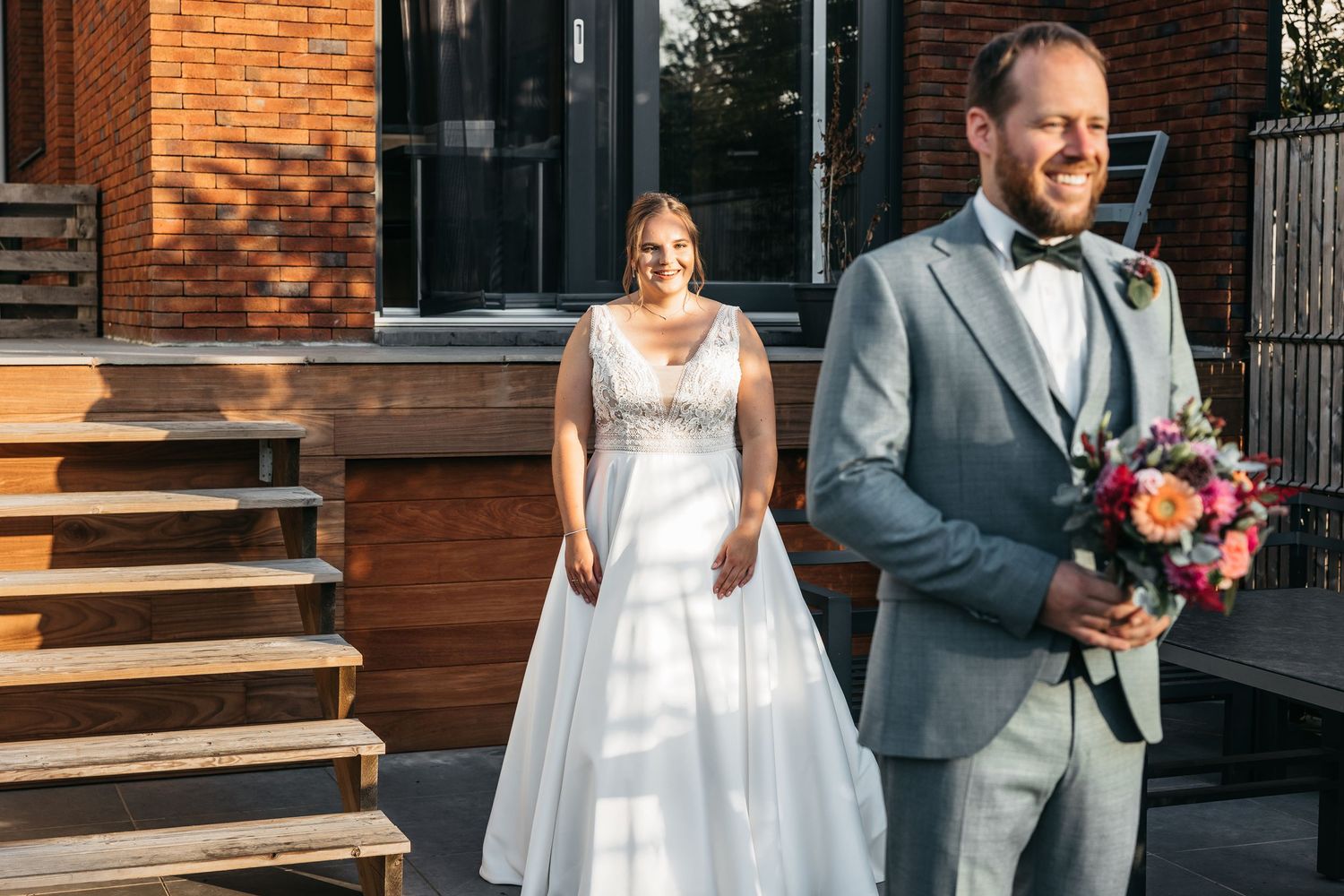 Bride in white gown smiles at groom in gray suit holding colorful bouquet during first look outside brick building.
