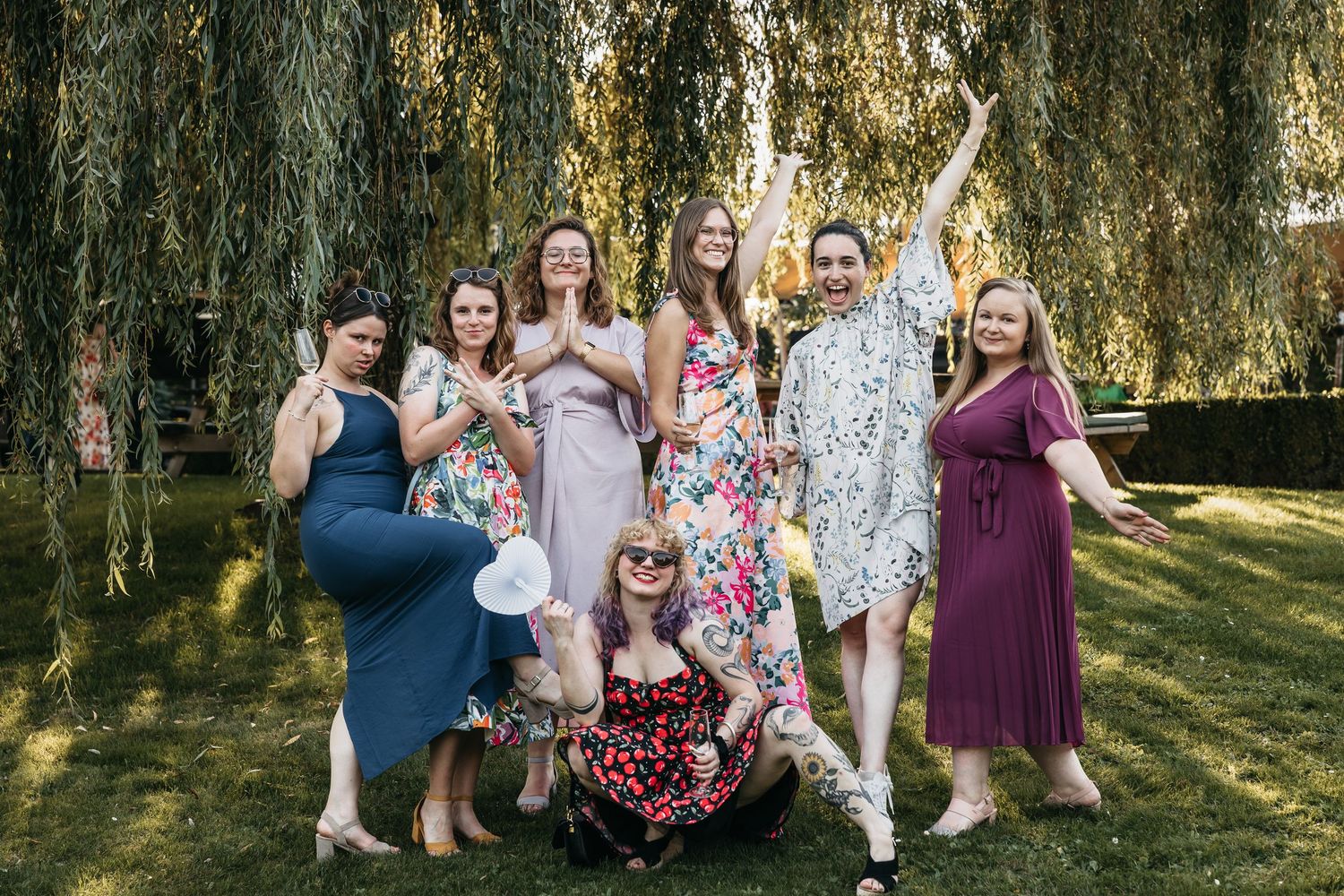 Group of female wedding guests laughing and posing playfully under a weeping willow tree.