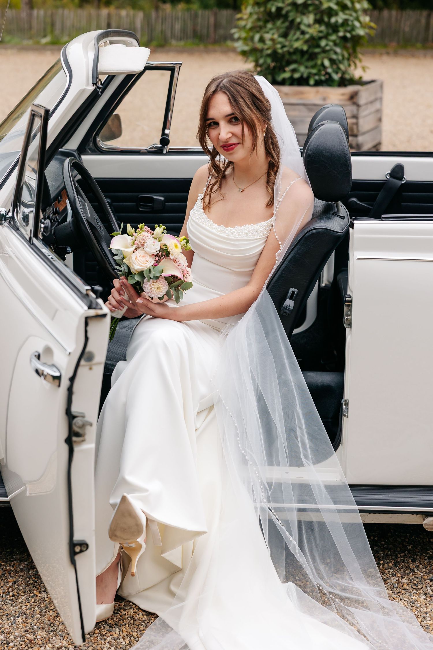 Smiling bride in white gown and veil seated in vintage white convertible car holding bouquet.