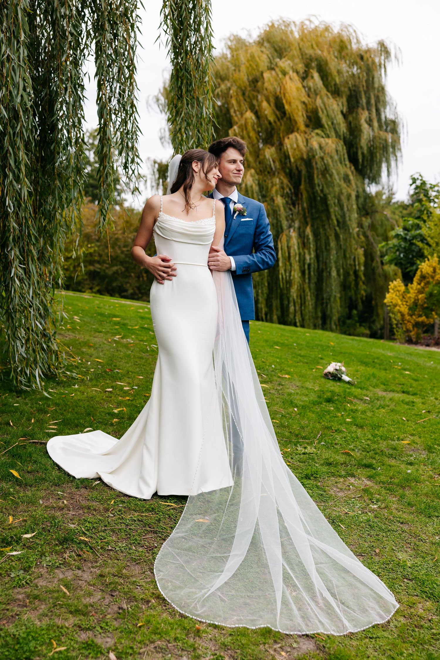 Elegant bride in fitted white gown with cathedral veil stands with groom in blue suit outdoors.