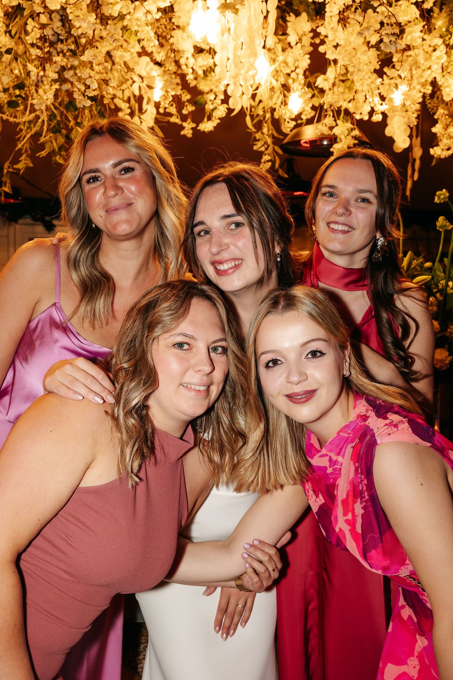 Five bridesmaids in pink and rose dresses smiling together under floral chandelier at wedding reception.