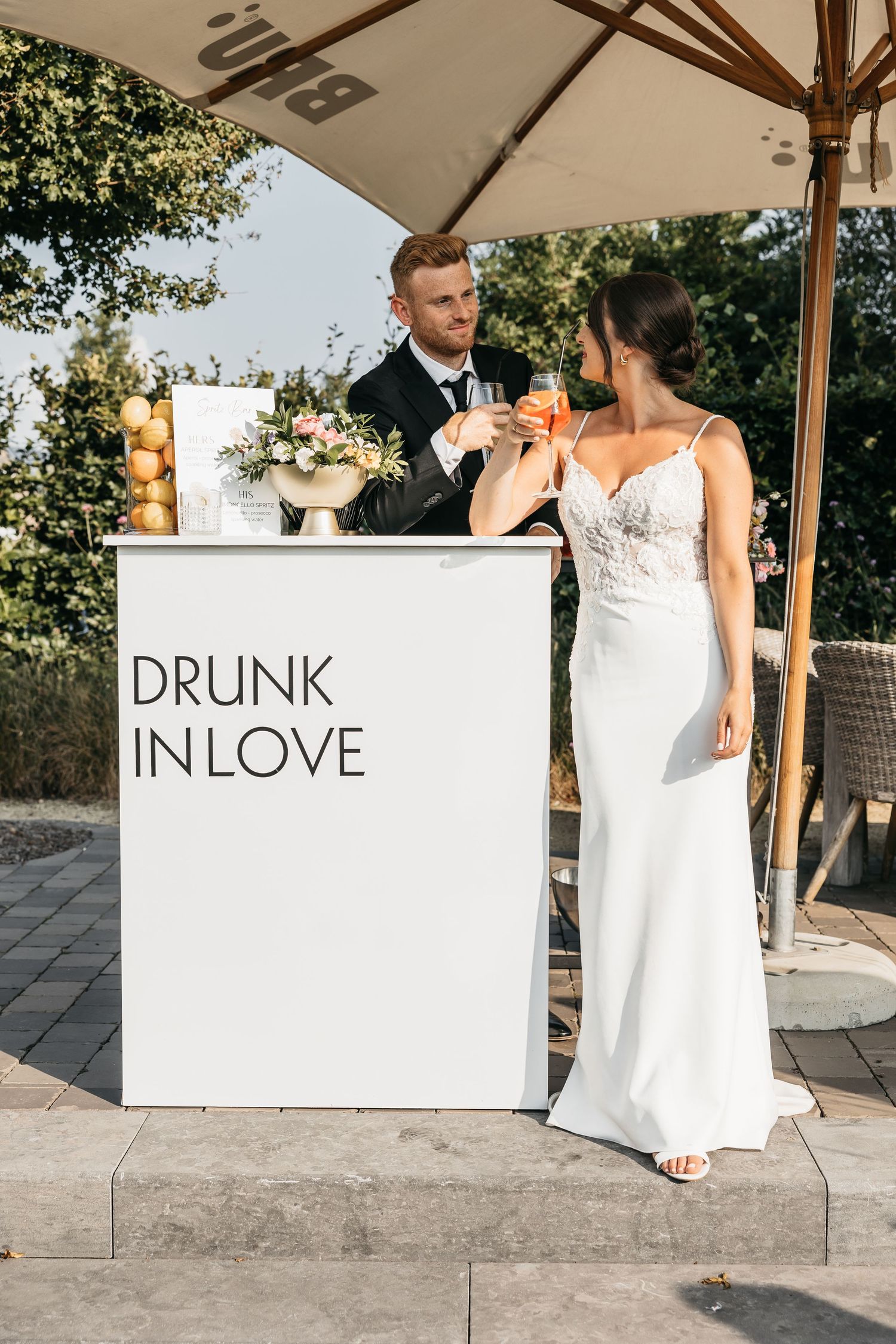 Bride and groom toasting champagne at a white bar sign reading Drunk In Love at outdoor wedding.