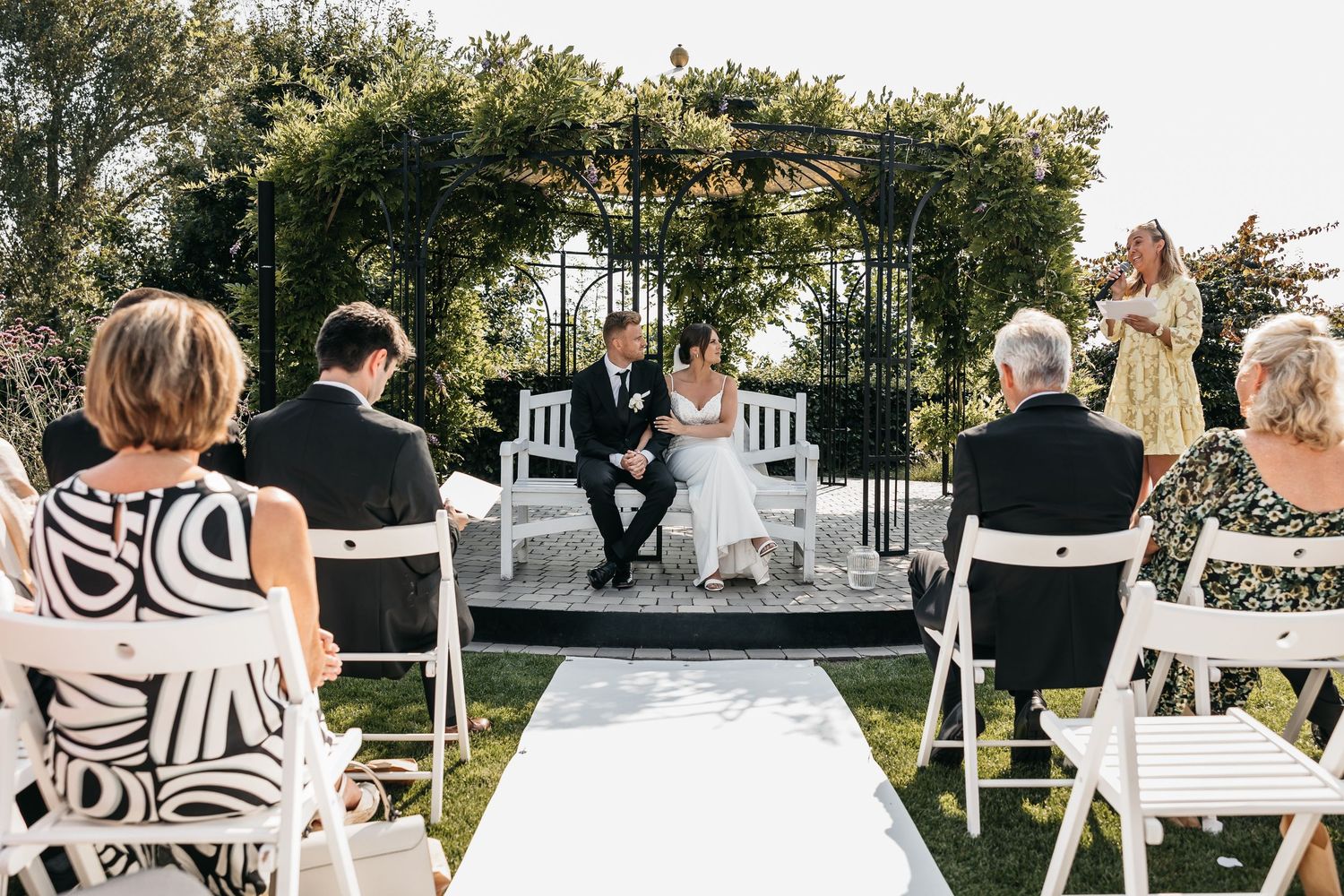 Elegant outdoor wedding ceremony with couple seated on white bench under black iron pergola in garden.