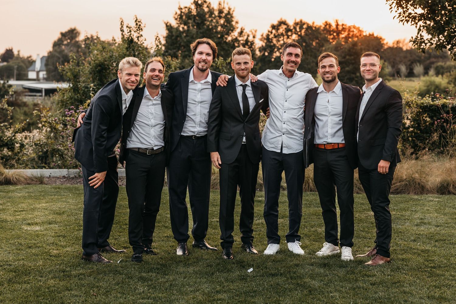 Groom with groomsmen in dark suits smiling together outdoors at golden hour wedding.