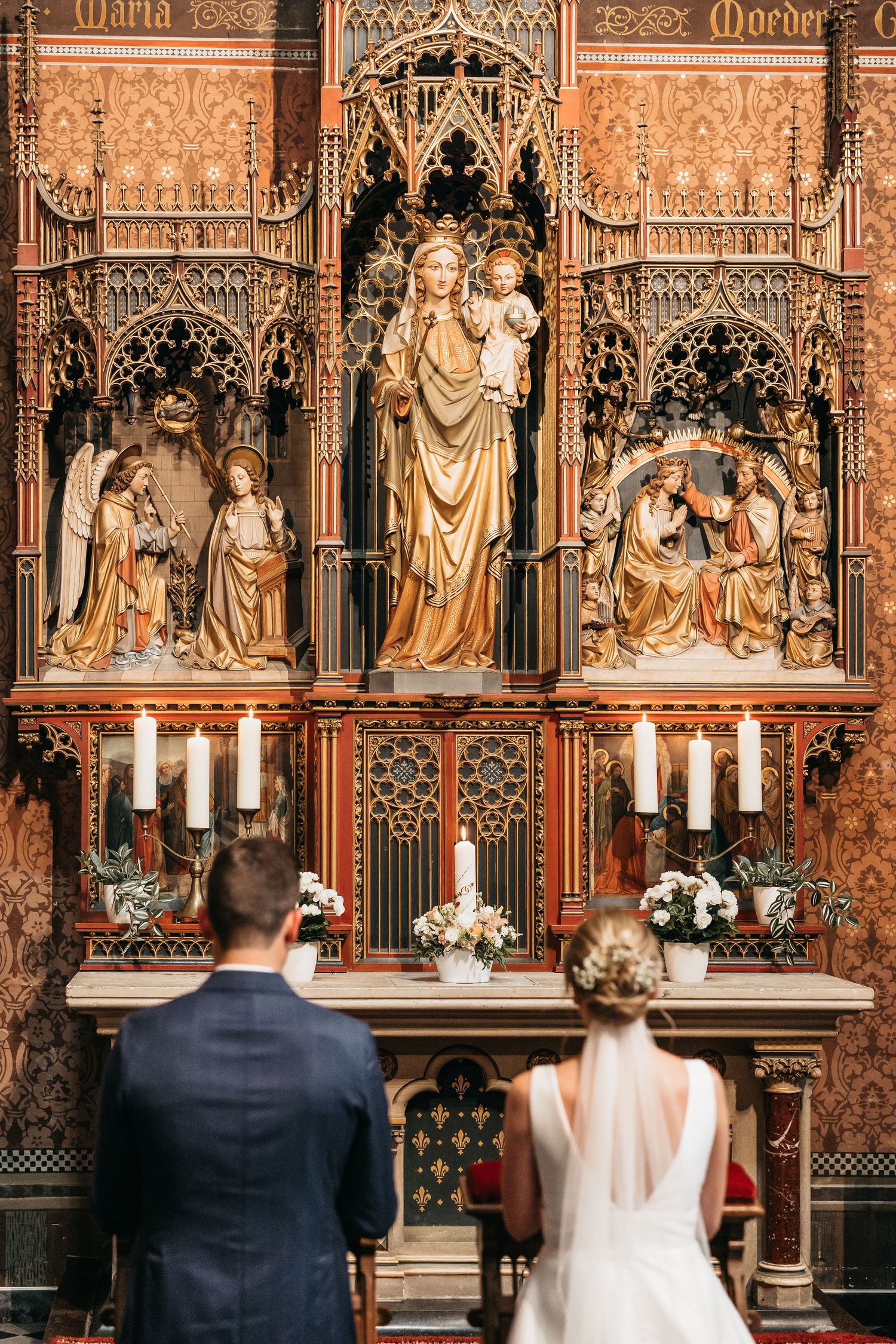Wedding couple standing before elaborate Gothic wooden altar with Virgin Mary statue and candles.