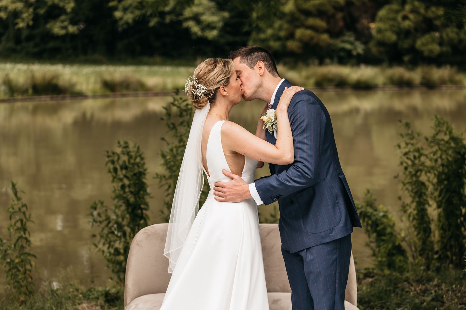 Bride and groom share first kiss beside a tranquil pond surrounded by lush greenery.