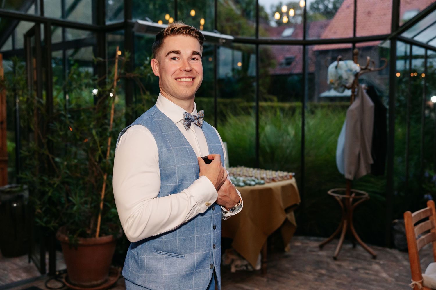 Handsome groom in blue checkered vest and bow tie smiling inside a greenhouse wedding venue.