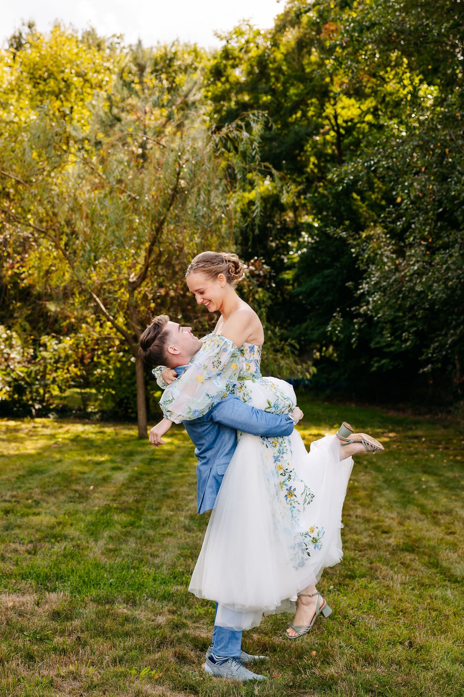 Groom in blue suit lifts smiling bride in floral wedding dress in a sunny garden setting.