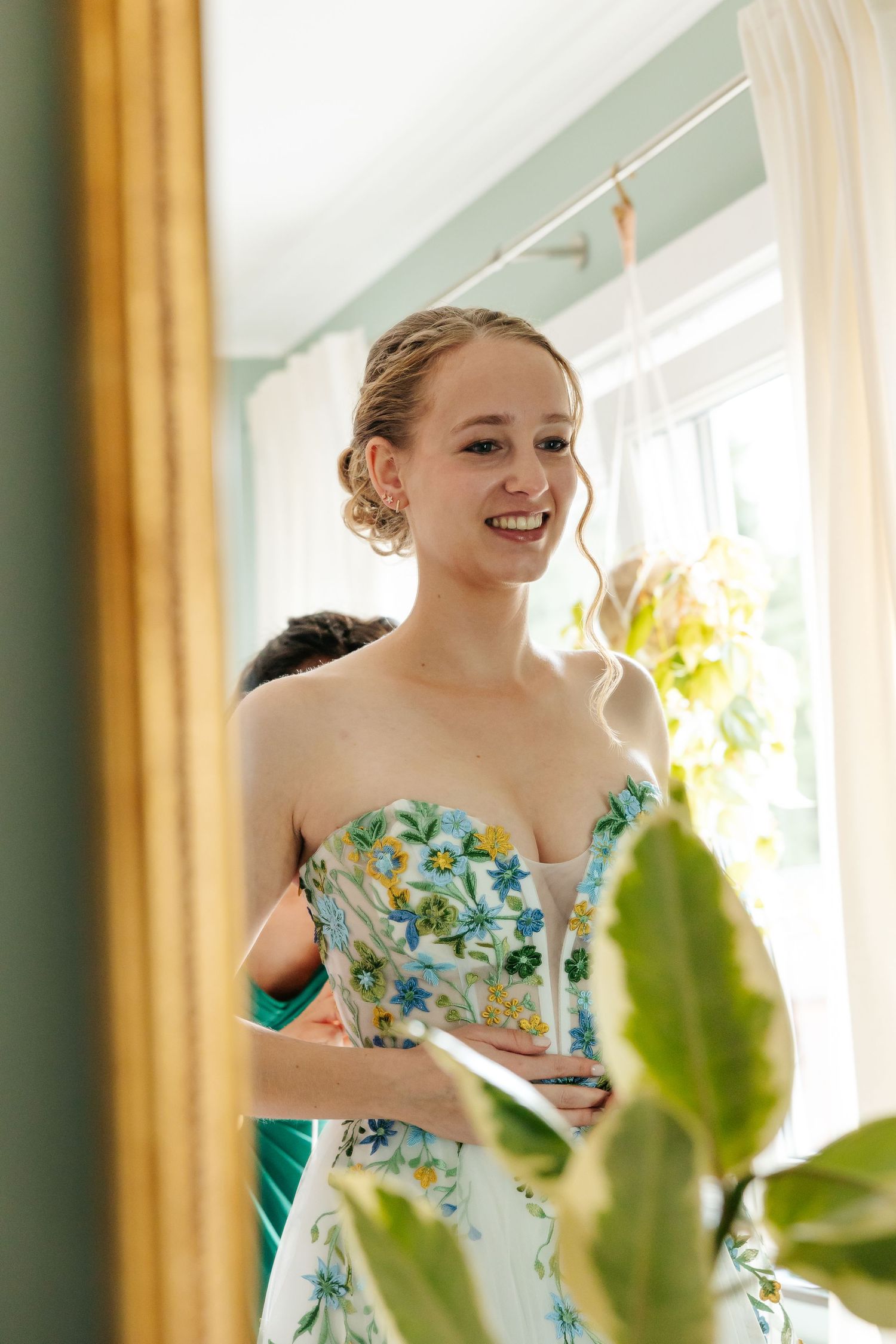 Happy bride in floral strapless wedding dress smiling while bridesmaid helps with final touches.