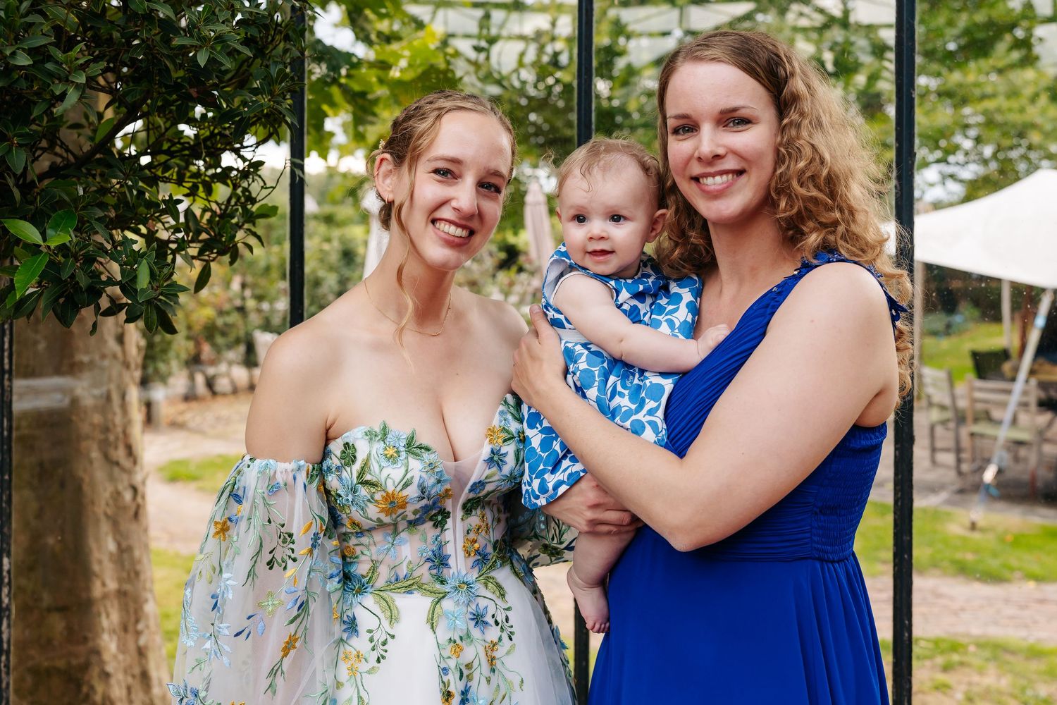 A bride in a floral dress and bridesmaid in blue smile with a baby at an outdoor wedding.