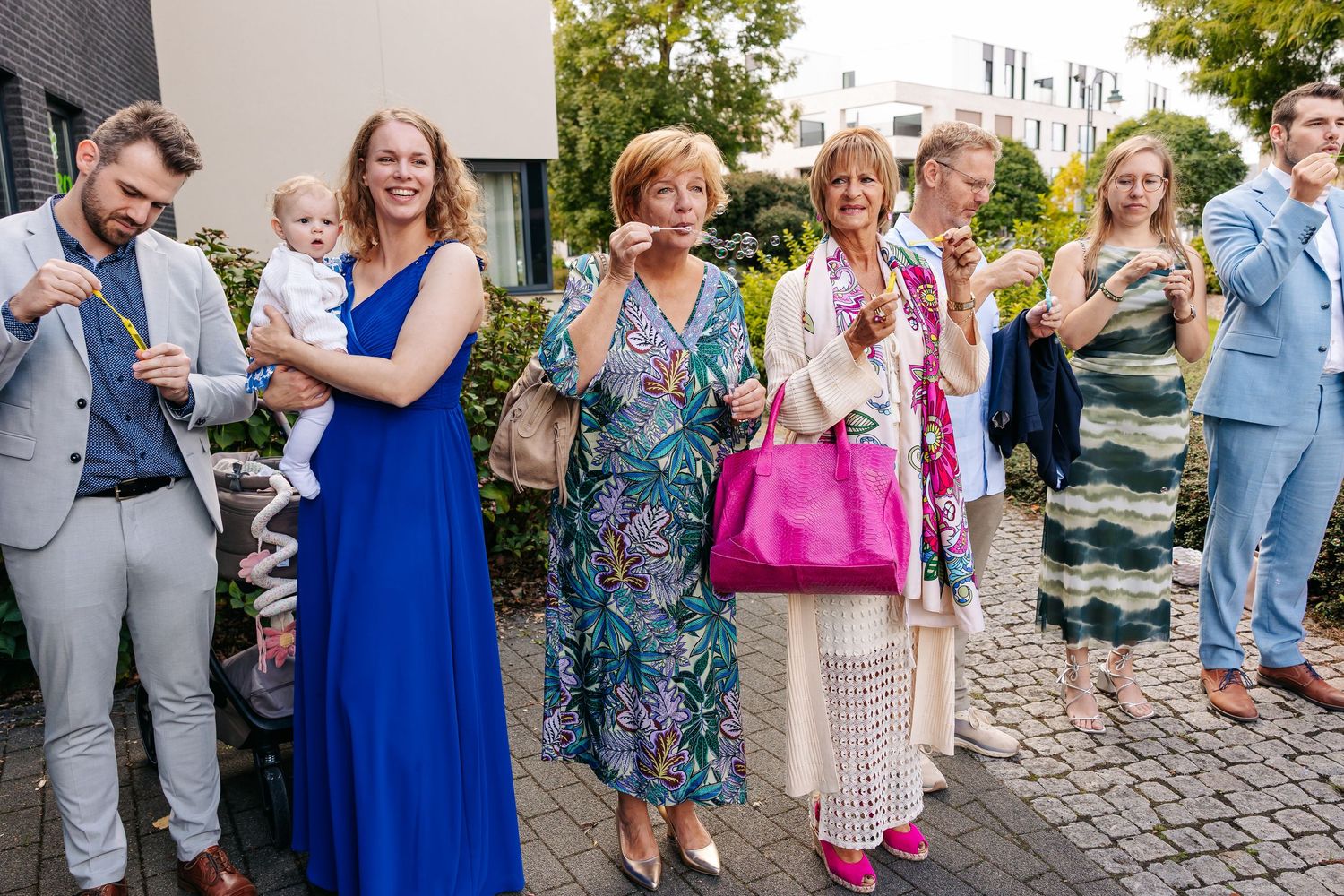 Smiling wedding guests in colorful outfits standing outside modern building during wedding celebration.