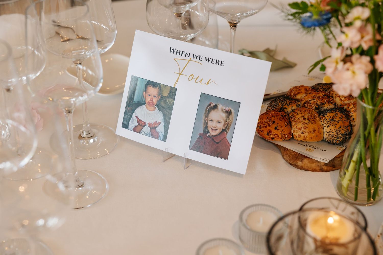 Wedding table number card displaying childhood photos with text When We Were Four beside bread and candles.