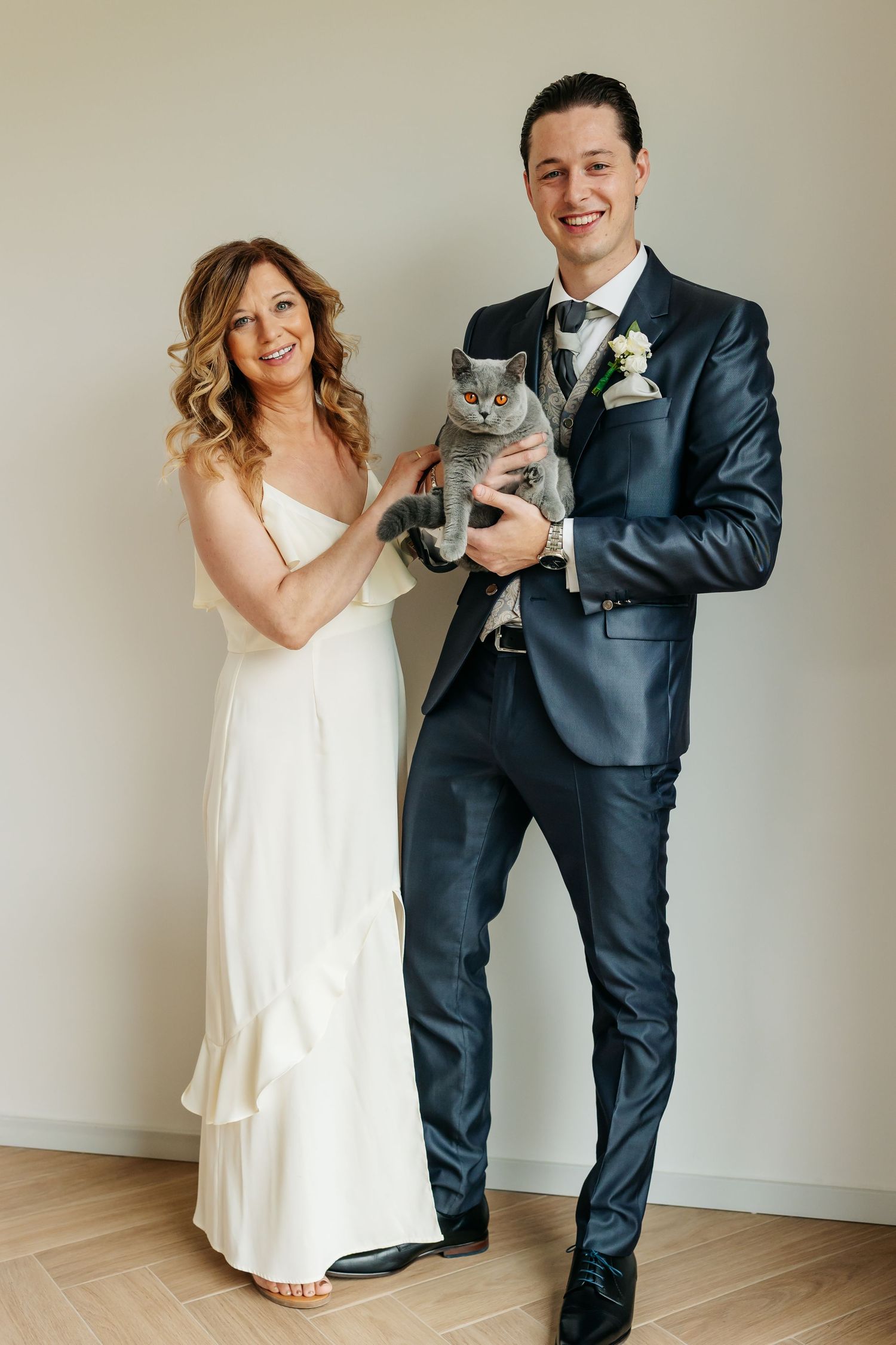 Smiling bride and groom in wedding attire pose together holding their gray British Shorthair cat.