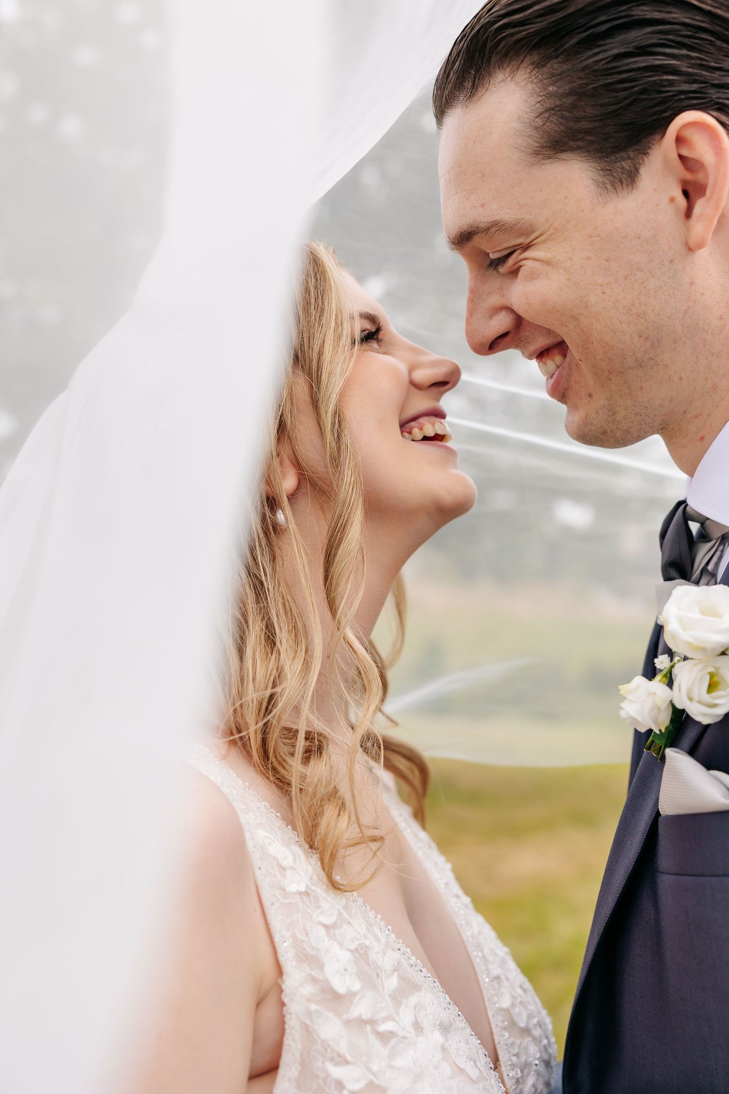 Bride and groom smile at each other under a billowing white veil on their wedding day outdoors.