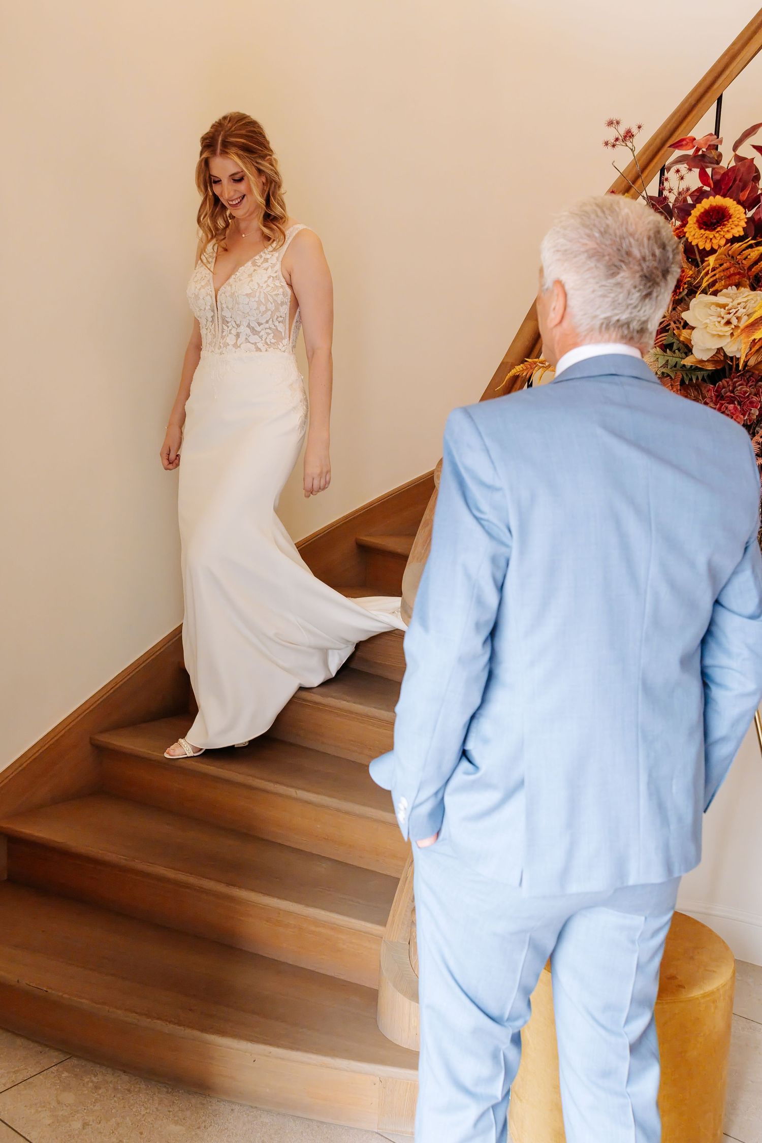 Bride in white lace gown descends wooden staircase as man in light blue suit watches below.