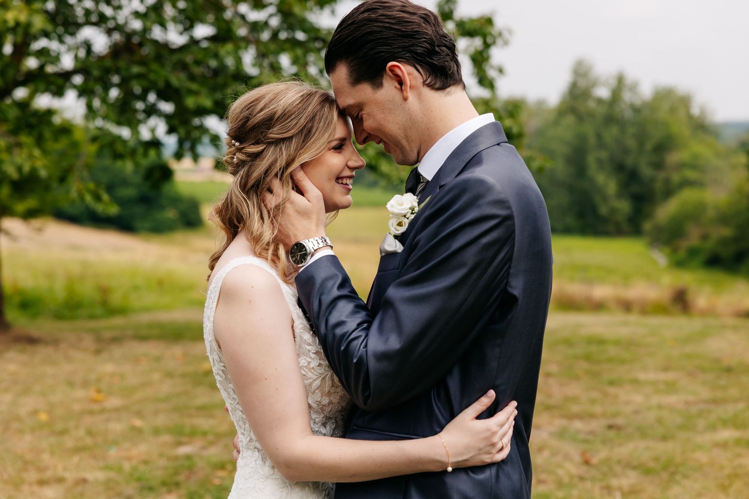 Bride and groom smiling and embracing closely outdoors on a sunny day with green fields behind them.