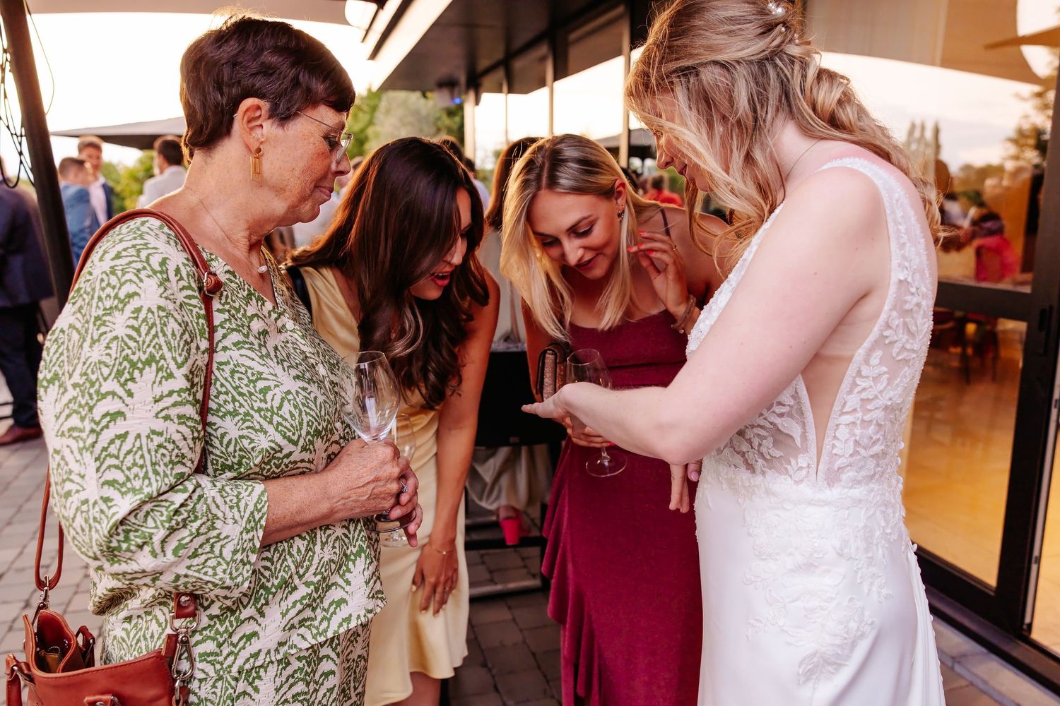 Bride in lace gown shares a moment with wedding guests looking at a phone outdoors.