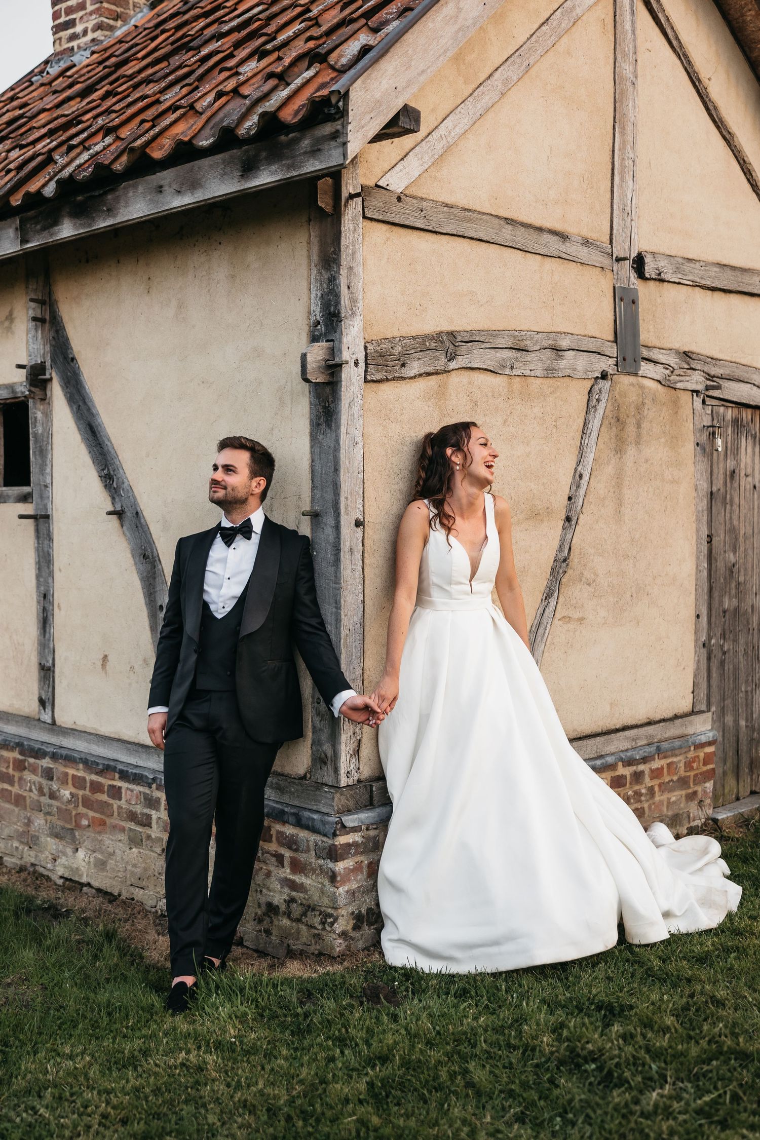 Bride and groom holding hands laughing beside a rustic timber-framed building on their wedding day.