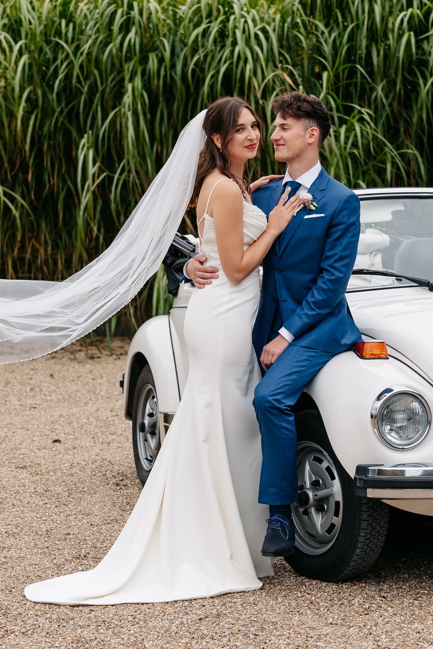 Bride and groom pose together leaning against a classic white Volkswagen Beetle.