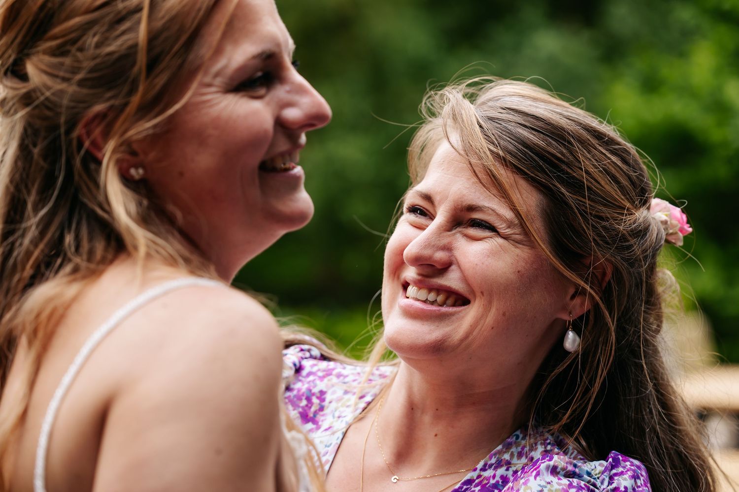 Two joyful women laughing together outdoors at a sunny wedding celebration.