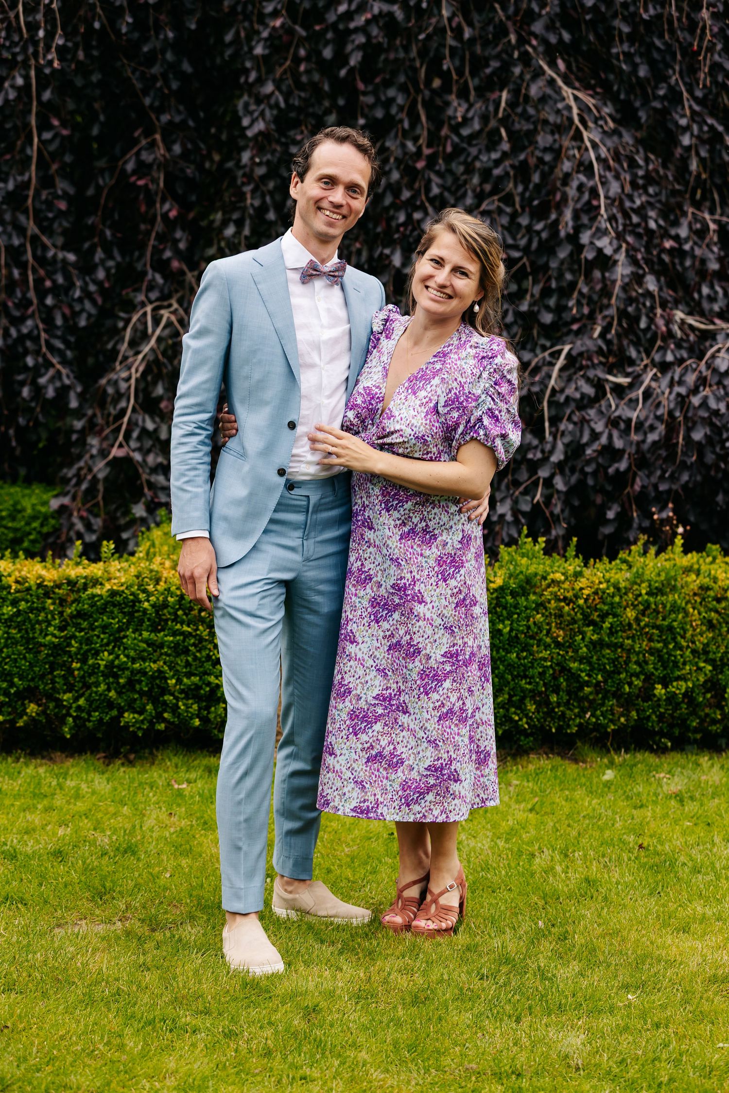 Smiling couple poses in garden, man in light blue suit and woman in purple floral dress.