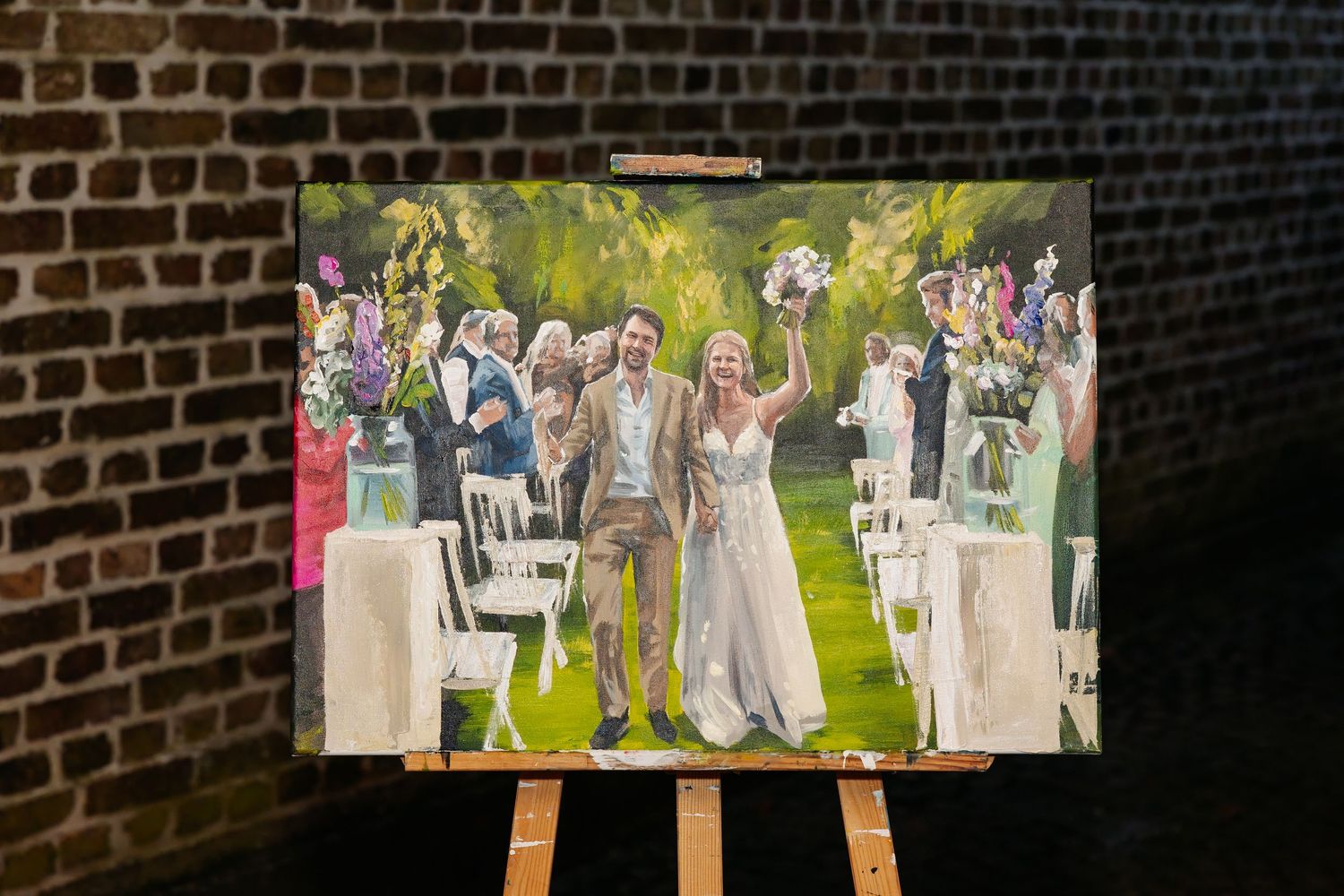 Wedding painting on easel displayed against brick wall showing bride and groom celebrating with guests.