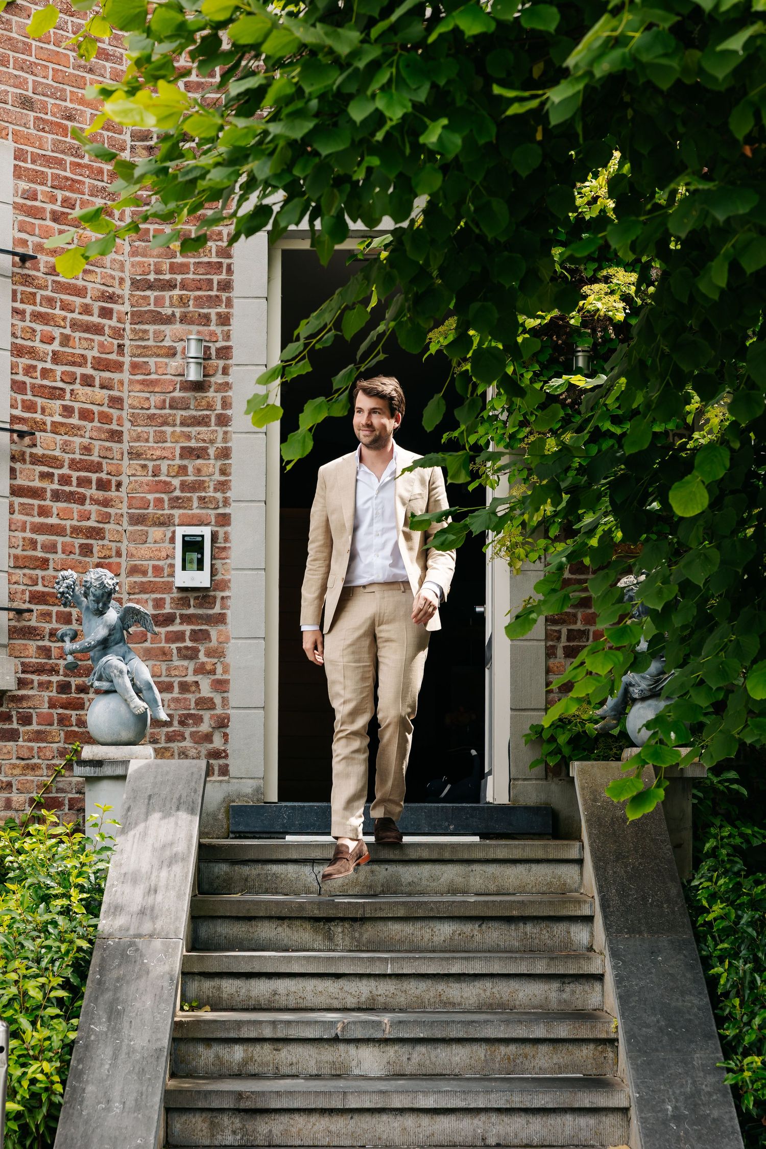 Groom in beige suit holding bouquet walking down stone steps of brick building entrance.