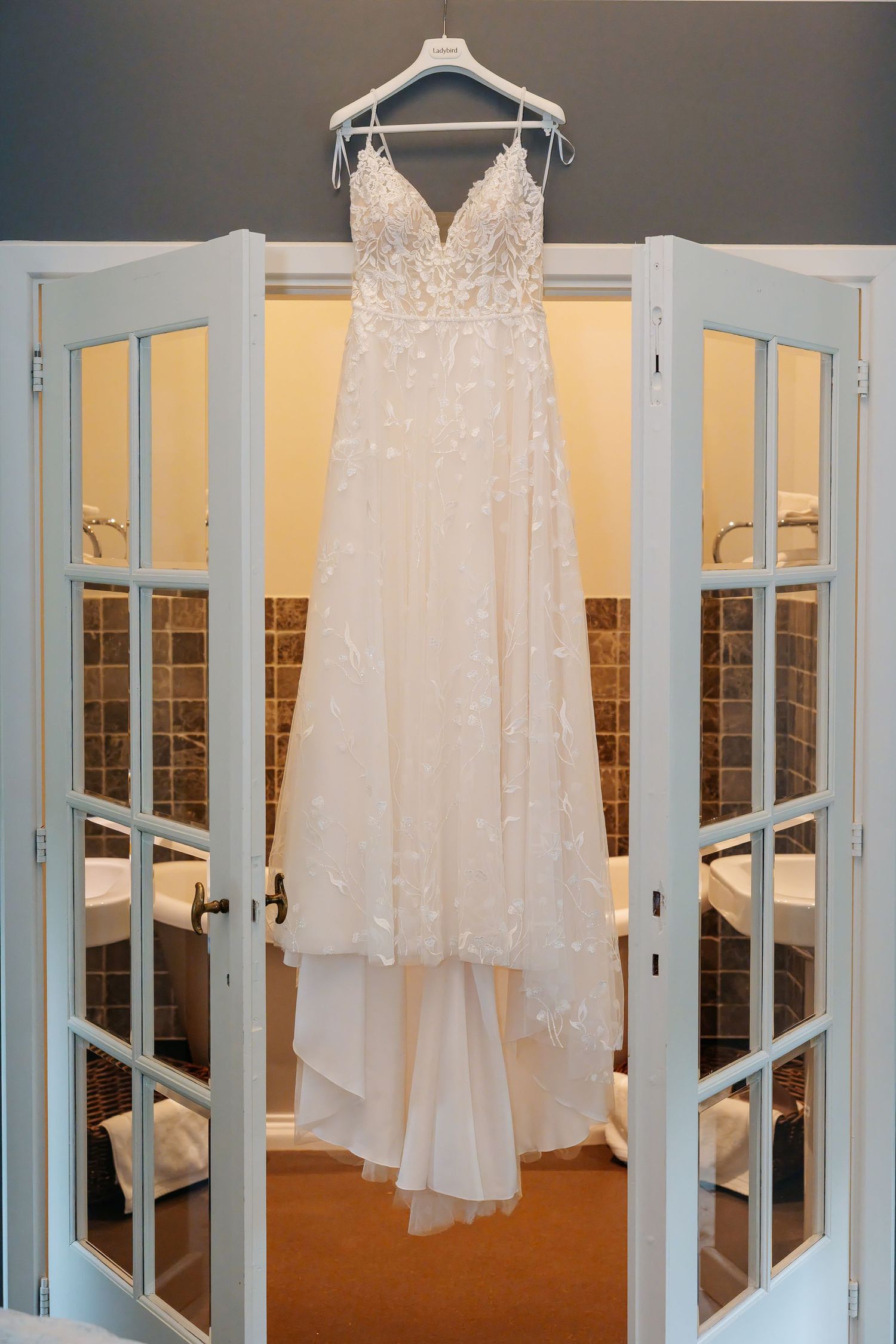 Elegant white lace A-line wedding dress hanging on French doors in a softly lit bathroom.