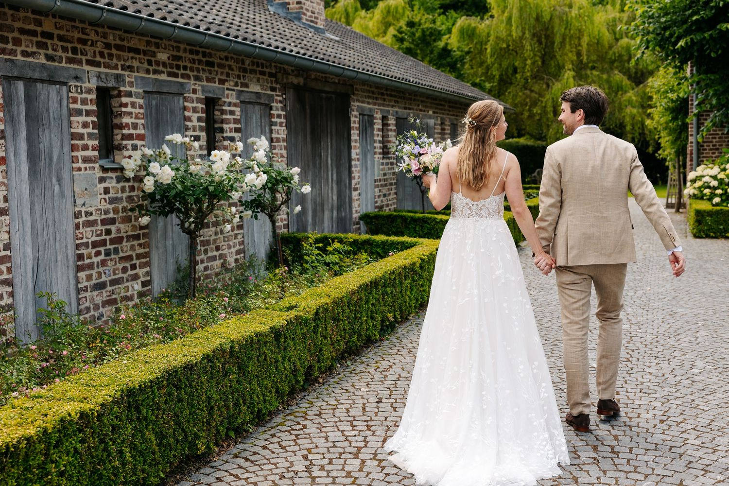 Bride and groom walking hand in hand along cobblestone path beside rustic brick wedding venue.