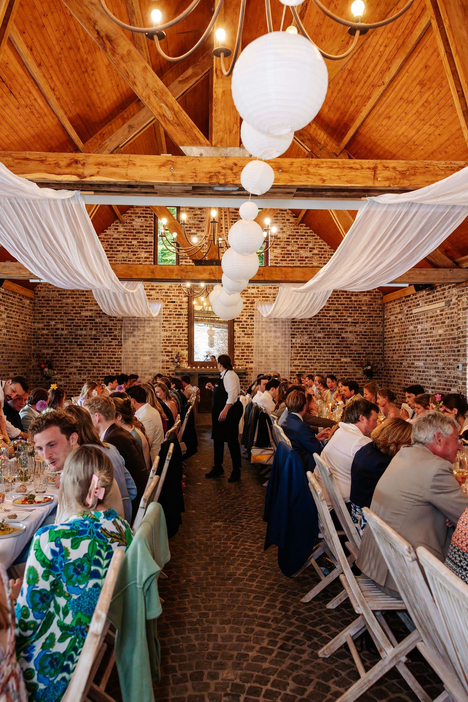 Wedding reception dinner in rustic brick barn venue with white lanterns, draped ceiling, and long guest tables.