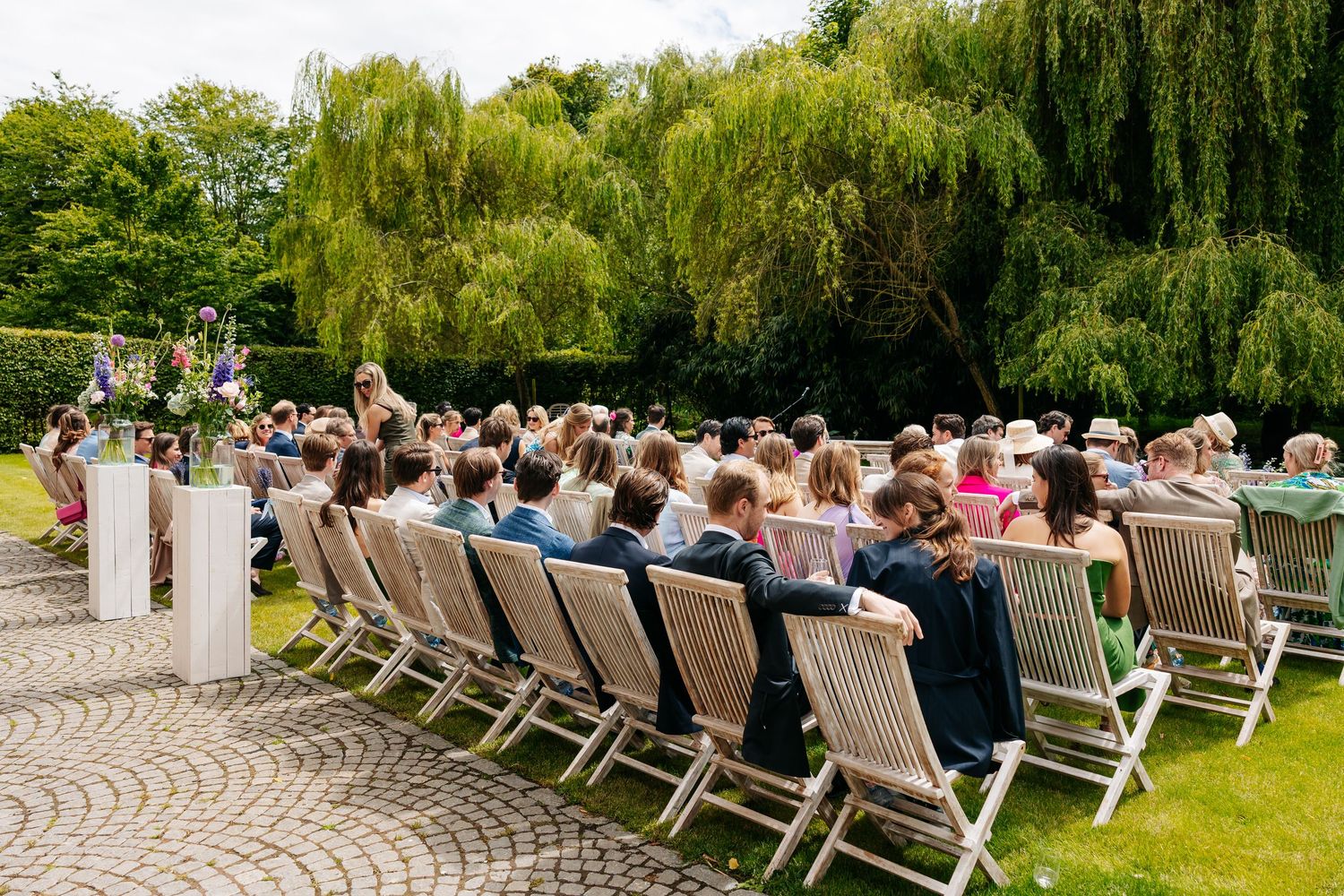 Outdoor wedding ceremony with guests seated in wooden chairs beneath a large weeping willow tree.
