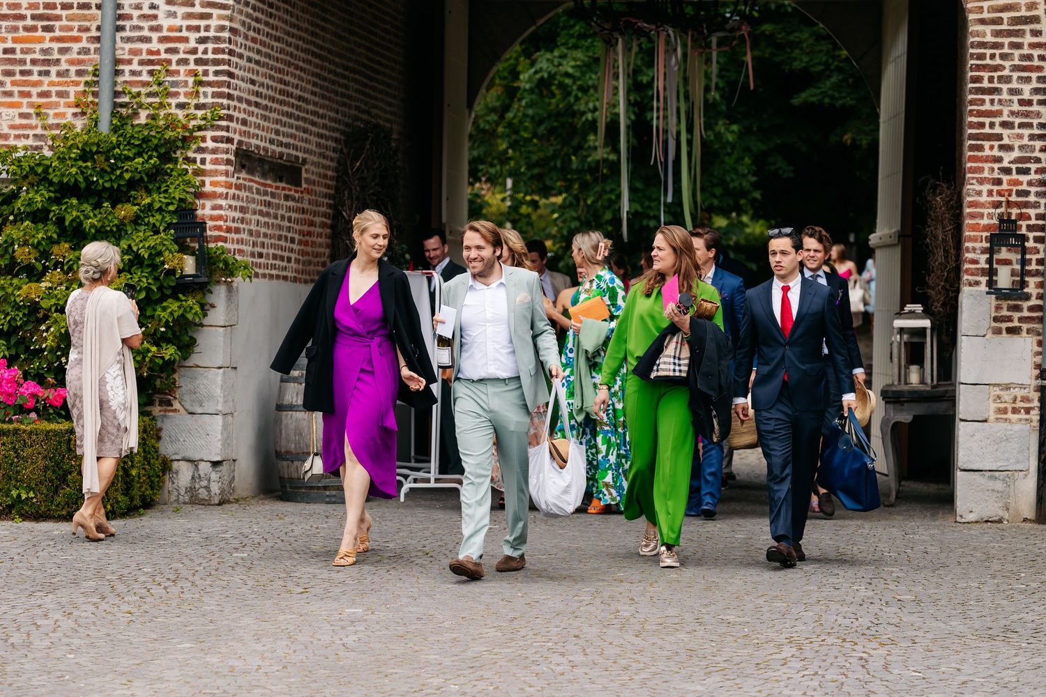 Colorfully dressed wedding guests walk together through a brick archway at an outdoor venue.