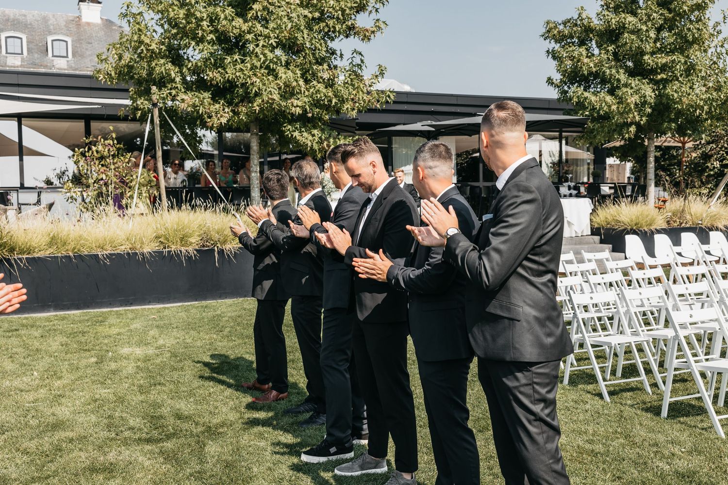 Groomsmen in black suits clapping and applauding beside rows of white chairs at outdoor wedding.