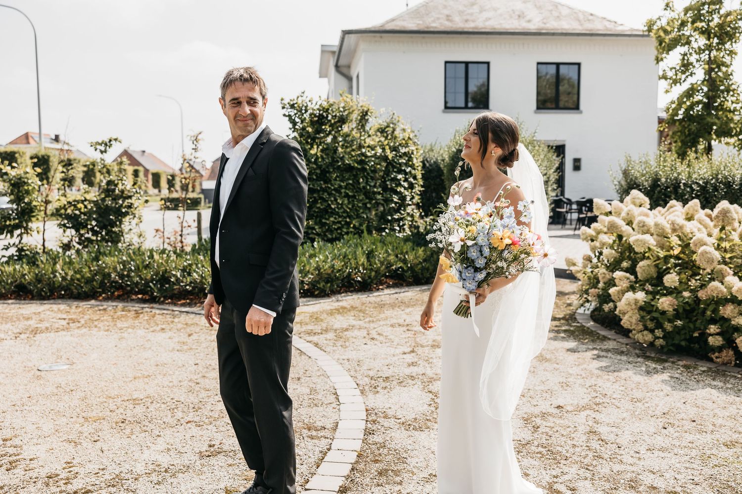 Groom in black tuxedo turns to see bride holding colorful bouquet during outdoor first look moment.
