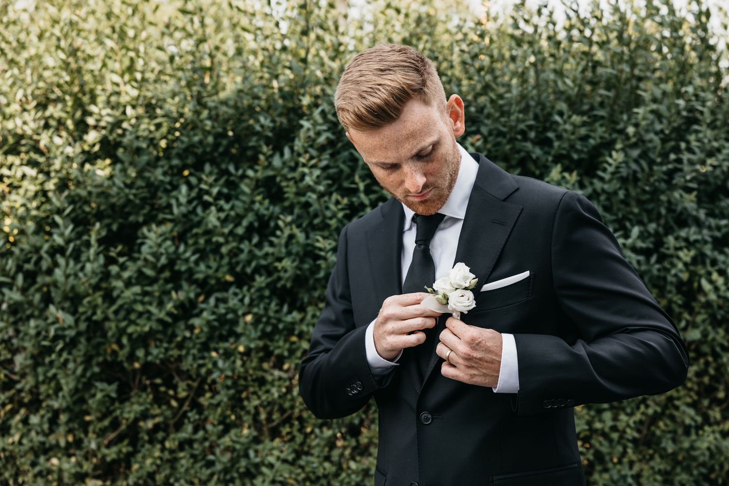 Groom in black suit carefully pinning white rose boutonniere to lapel in front of green hedge.