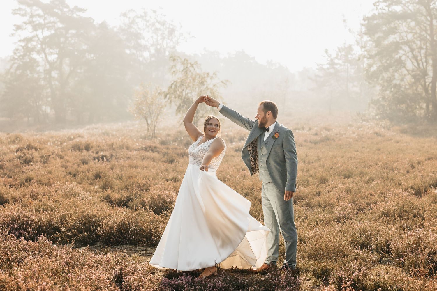 Smiling bride twirling in white gown as groom spins her in a sunlit heather meadow.