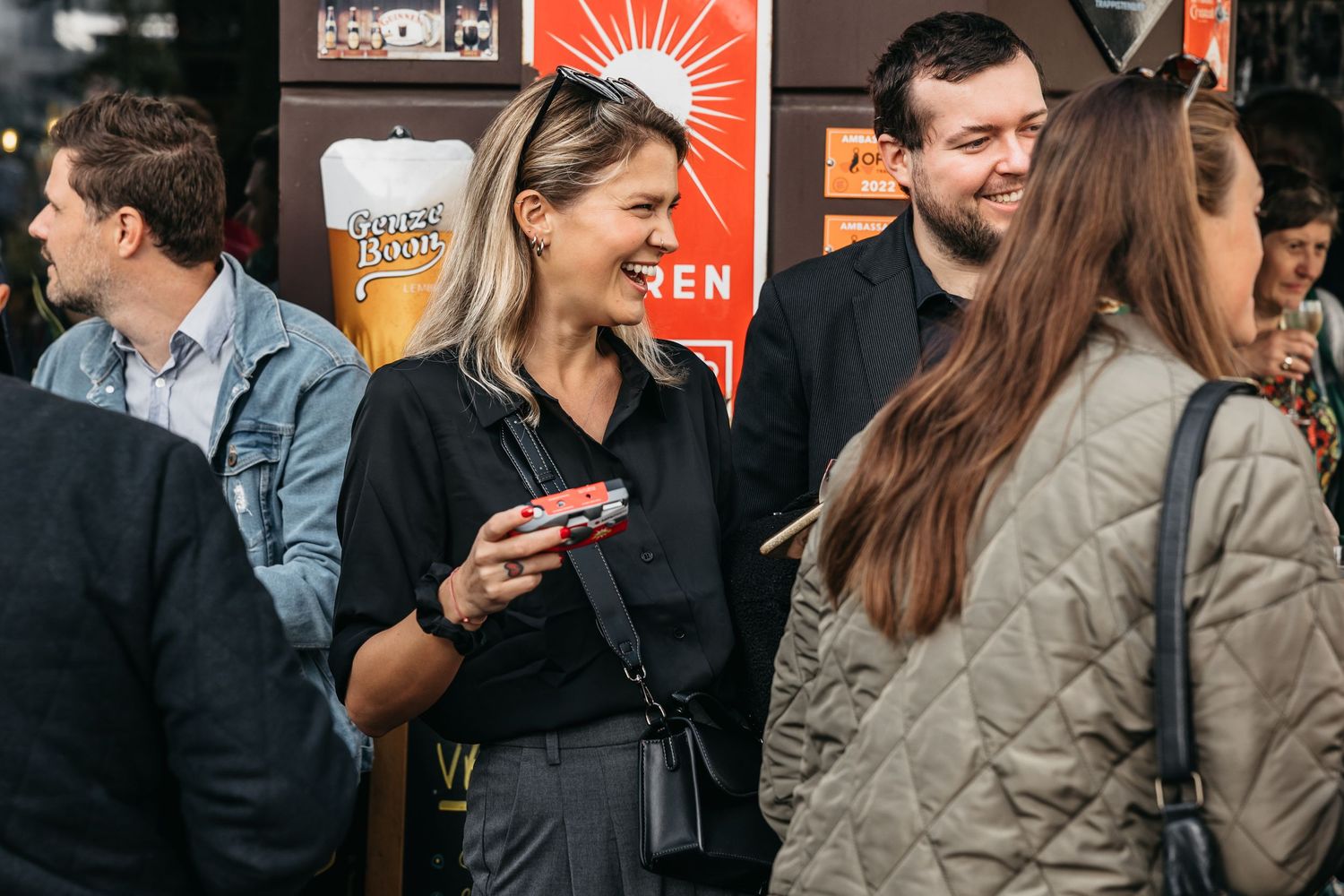 Smiling woman holding camera chatting with guests outside bar at lively evening wedding reception.