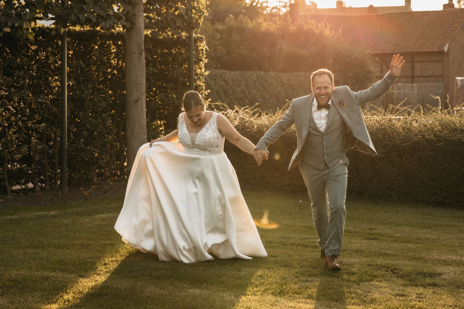 Bride and groom joyfully running hand in hand across lawn in golden sunset light.