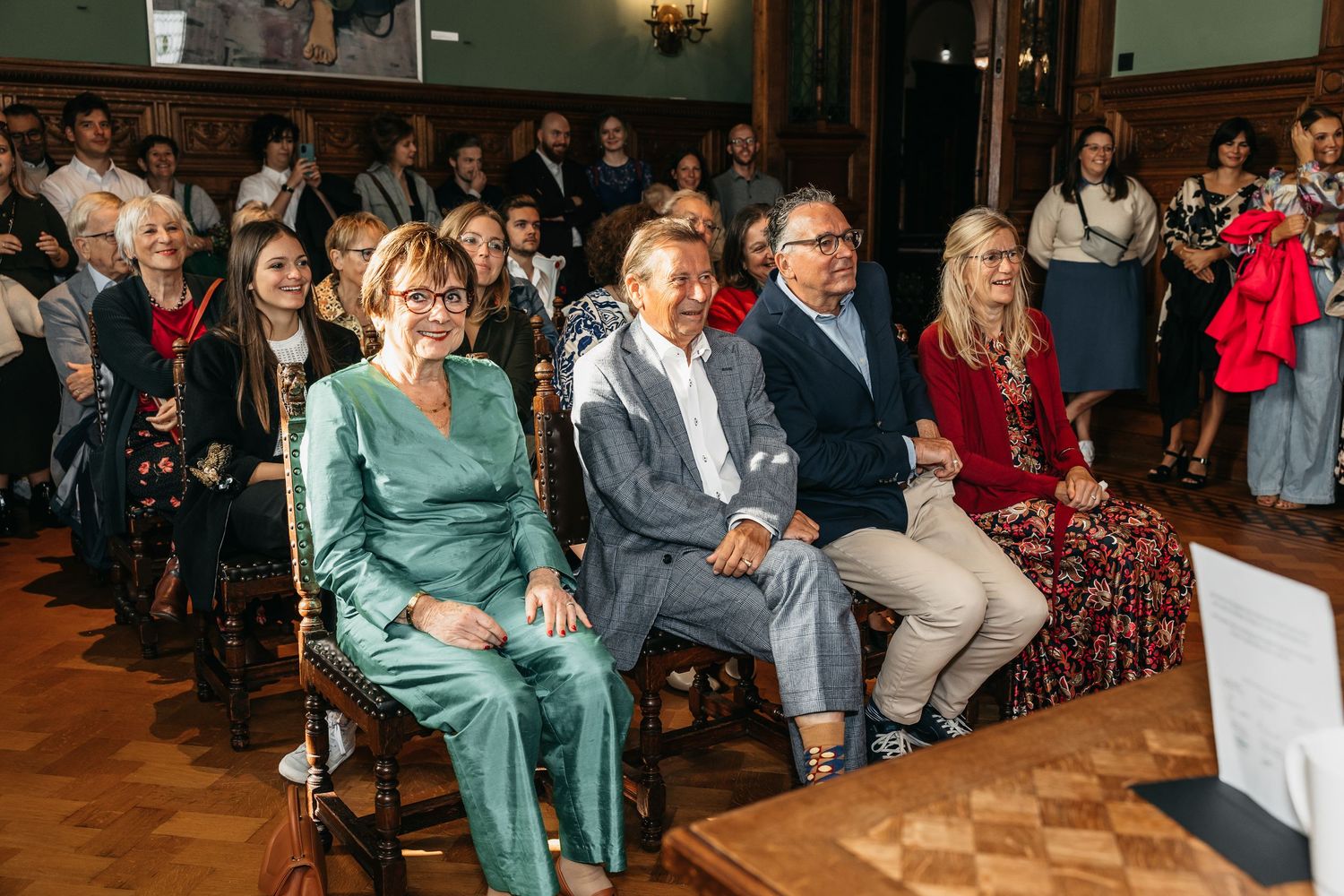 Smiling wedding guests in formal attire seated in rows at an elegant wood-paneled ceremony hall.