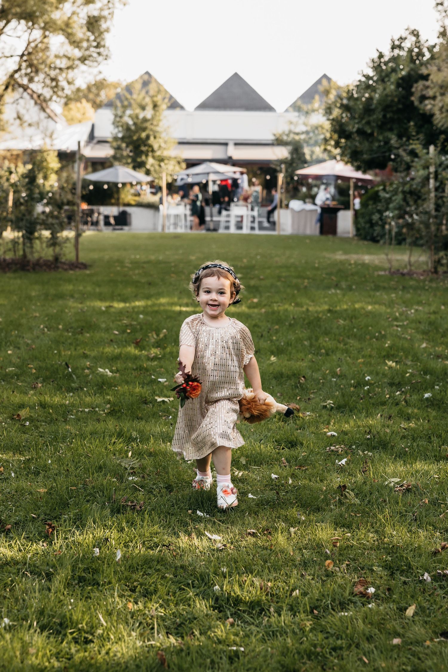 Adorable flower girl in cream dress running across green lawn holding flowers at outdoor wedding reception.