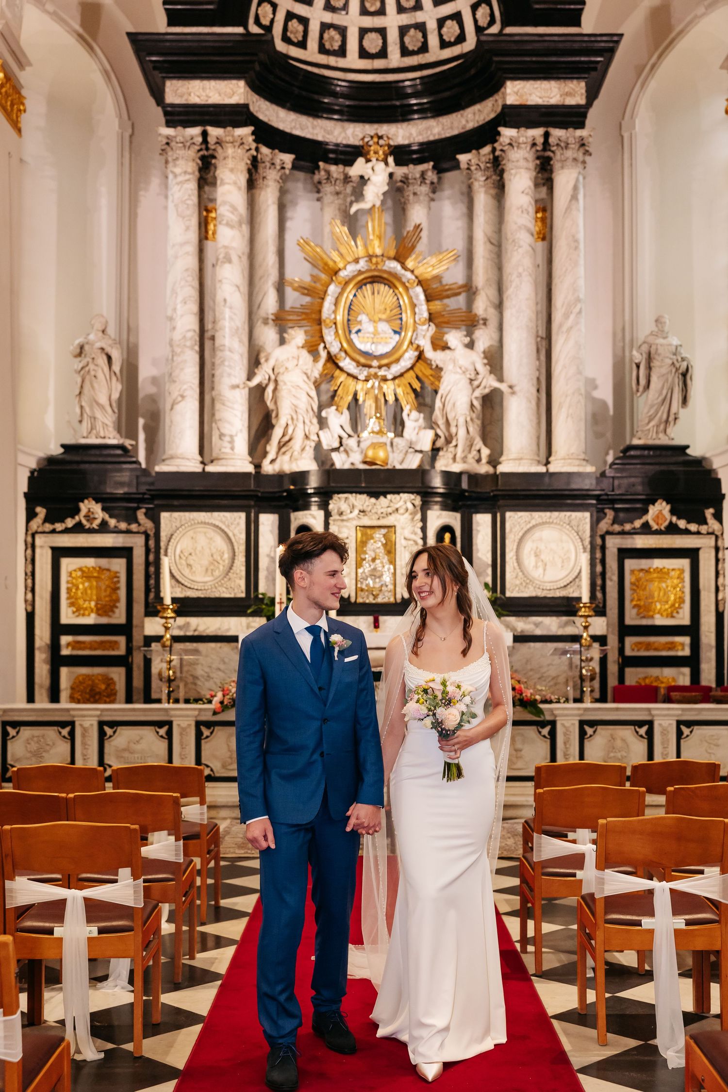 Newlyweds walk hand in hand down red aisle before grand baroque church altar.