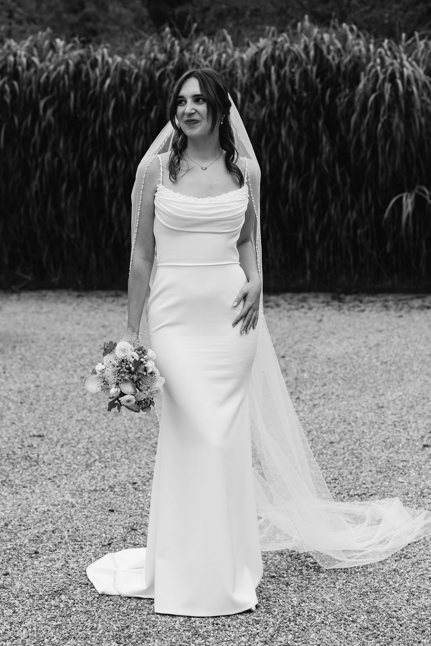 Black and white portrait of bride in fitted white gown and long veil holding floral bouquet.