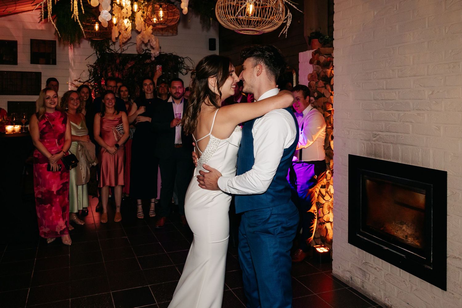 Bride and groom share intimate first dance embrace near fireplace with wedding guests watching in warm lighting.